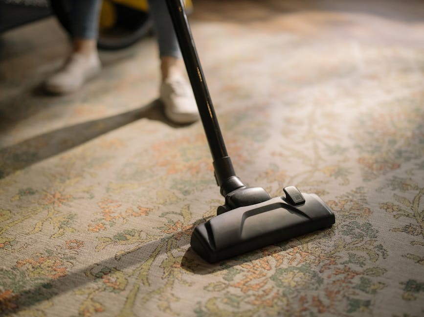 A woman with curly hair, dressed in a beige jacket and dark jeans, is using a yellow vacuum cleaner with a black hose to clean a patterned carpet in a living room. The room has wooden flooring adjacen