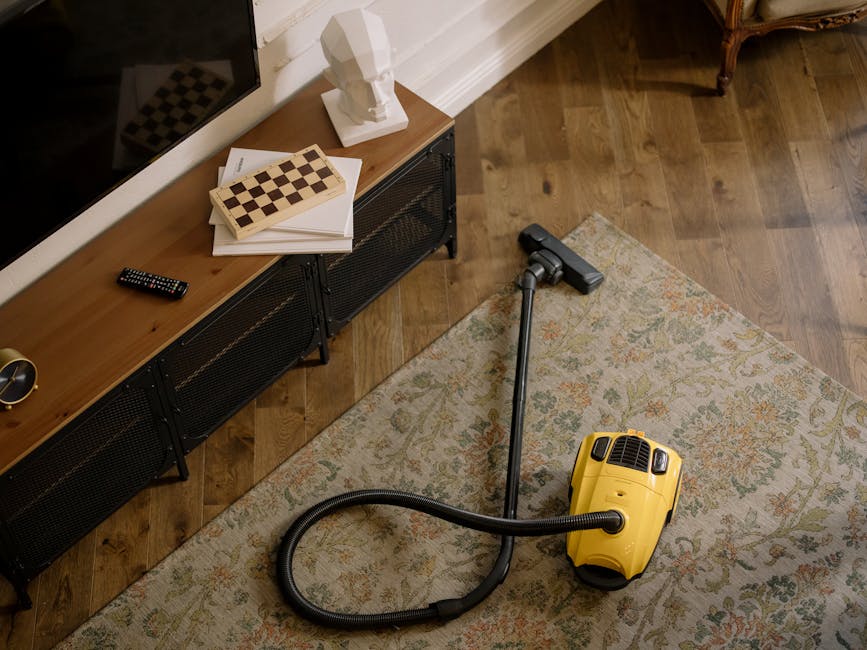 A woman using a yellow vacuum cleaner with a hose attachment to perform deep surface cleaning on a patterned area rug in a living room. The room has hardwood flooring visible at the edges, with a leat