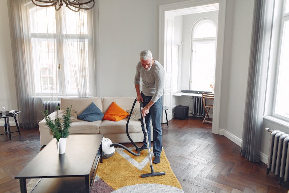 Close-up of a person vacuuming a patterned area rug with a black vacuum cleaner, highlighting the deep cleaning process. The rug features intricate floral designs in soft pastel shades, laid on a hard