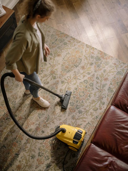 A person using a yellow vacuum cleaner to deep clean a patterned area rug on a wooden floor in a living room. The individual is wearing a beige jacket, blue jeans, and white sneakers, focusing on surface cleaning around the edge of a brown leather sofa. The room features natural lighting, highlighting the cleanliness of the rug and surrounding surfaces, with visible wooden flooring and part of an electronic device near the carpet. Hornchurch Carpet Cleaners specializes in professional domestic cleaning services, ensuring hygienic and well-maintained environments.