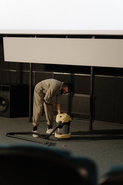 A person dressed in a light-colored jumpsuit is performing deep cleaning on a carpeted floor using a vacuum cleaner with a handheld attachment. The setting appears to be an indoor space with dark walls, a radiator, and a large white screen or panel mounted on the wall above the individual. The room is well-lit, highlighting the cleanliness of the carpet and surrounding surfaces. Visible equipment, including a black speaker or subwoofer, is positioned to the side, and the overall environment suggests a professional cleaning operation focused on surface cleaning and sanitisation. The image underscores the expertise of Hornchurch Carpet Cleaners in providing thorough deep cleaning services in commercial or public spaces.