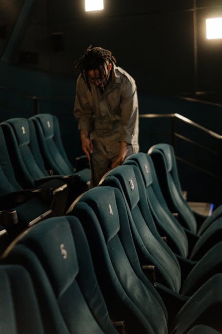 A person using a yellow vacuum cleaner to deep clean a patterned area rug on a wooden floor in a living room. The individual is wearing a beige jacket, blue jeans, and white sneakers, focusing on surface cleaning around the edge of a brown leather sofa. The room features natural lighting, highlighting the cleanliness of the rug and surrounding surfaces, with visible wooden flooring and part of an electronic device near the carpet. Hornchurch Carpet Cleaners specializes in professional domestic cleaning services, ensuring hygienic and well-maintained environments.